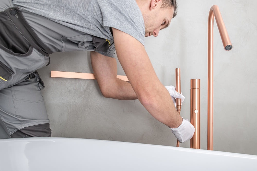 Bathroom fitter installing a modern copper freestanding bath tap next to a white tub.