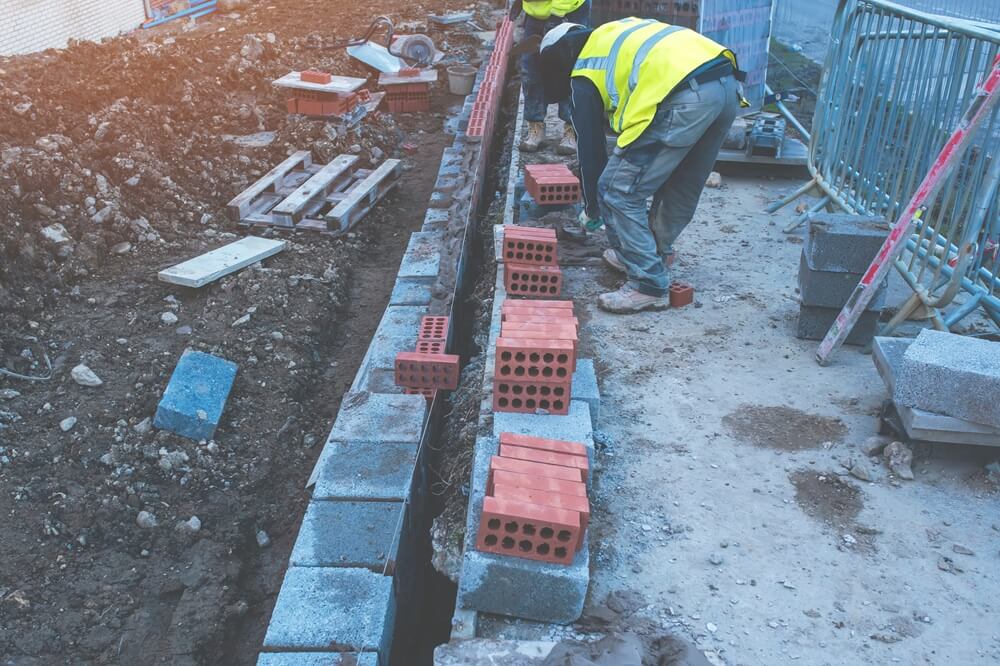 Bricklayers laying the first course of red bricks on a building site over concrete foundations