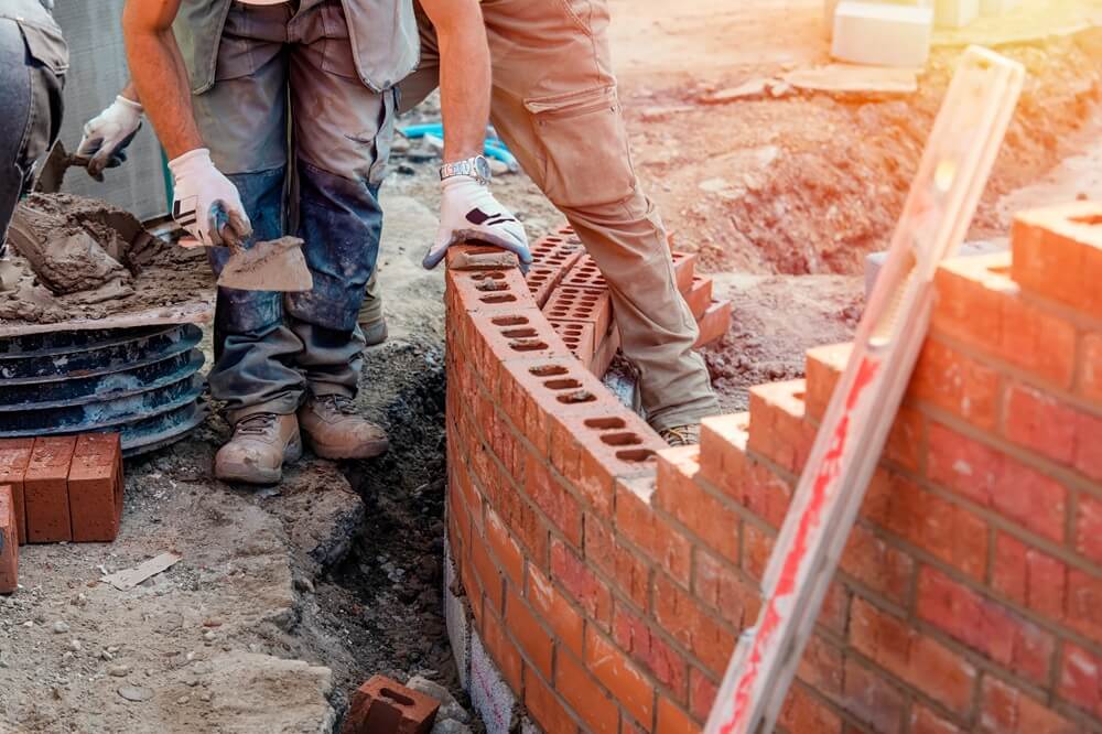 Two bricklayers constructing a curved red brick wall on a building site with tools and mortar