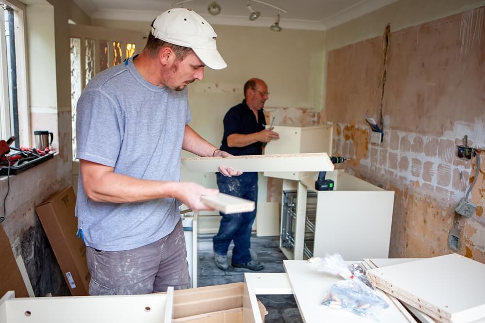 Two kitchen fitters assembling base units during a full kitchen renovation in a stripped plastered room