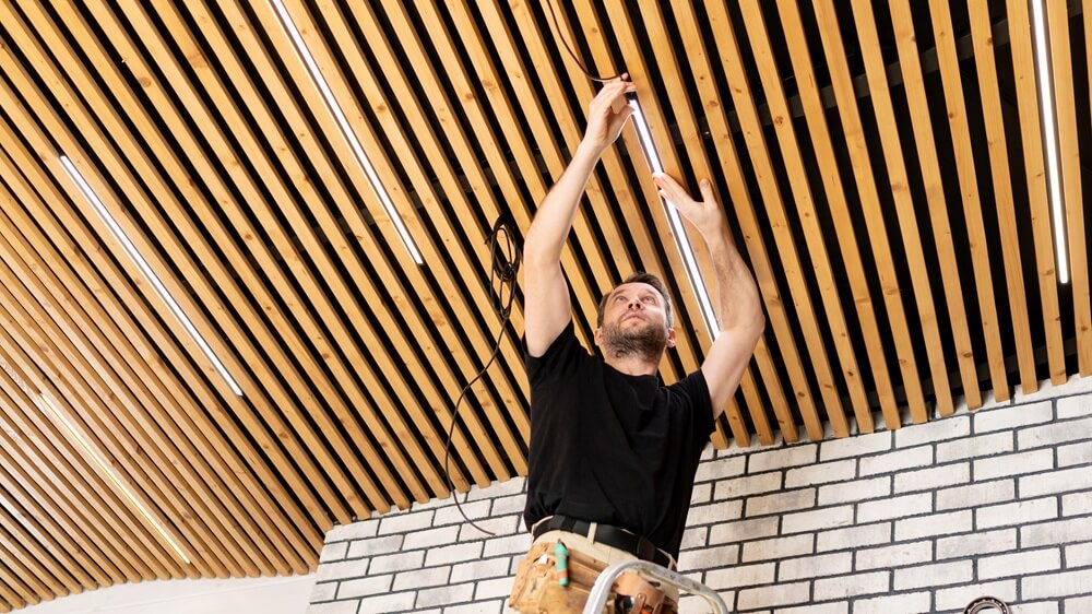 Electrician on ladder installing LED lighting in a suspended wooden ceiling inside a commercial space.