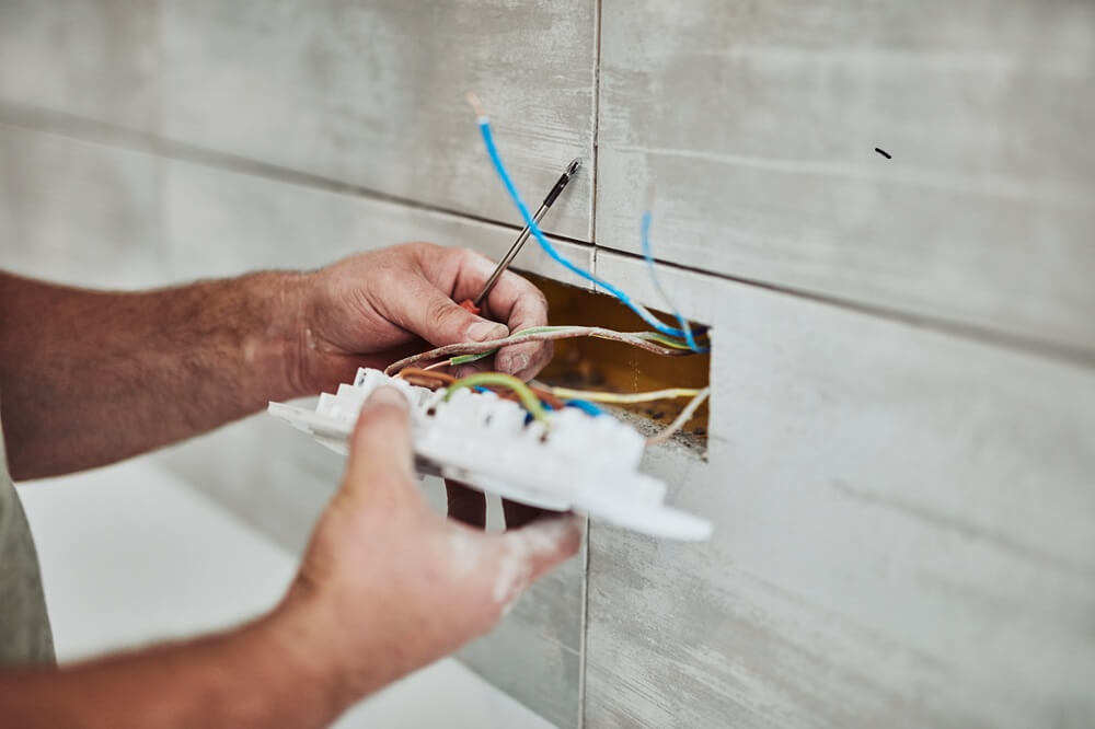 Electrician wiring live cables into a wall socket during installation in a tiled room.
