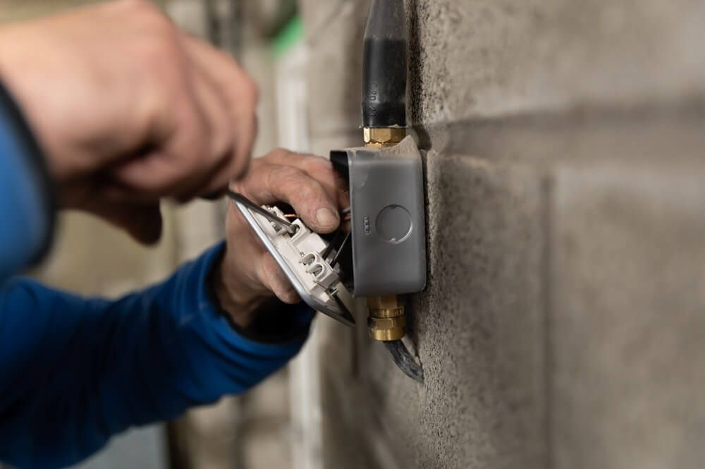 Electrician wiring a wall-mounted socket on a grey block wall during installation.