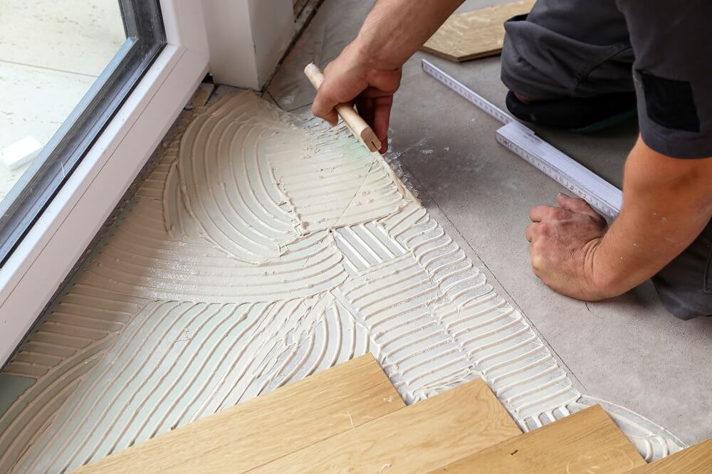 Flooring contractor spreading adhesive with a notched trowel before laying wooden floorboards.