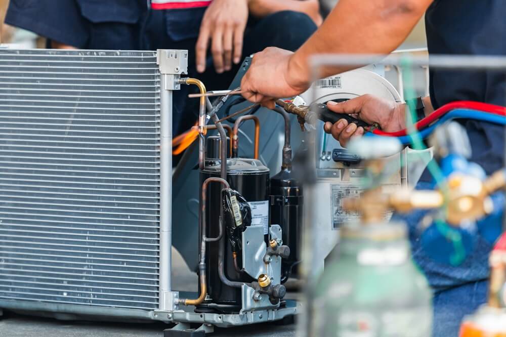 Engineer brazing copper pipework on an outdoor HVACR condensing unit during field maintenance.