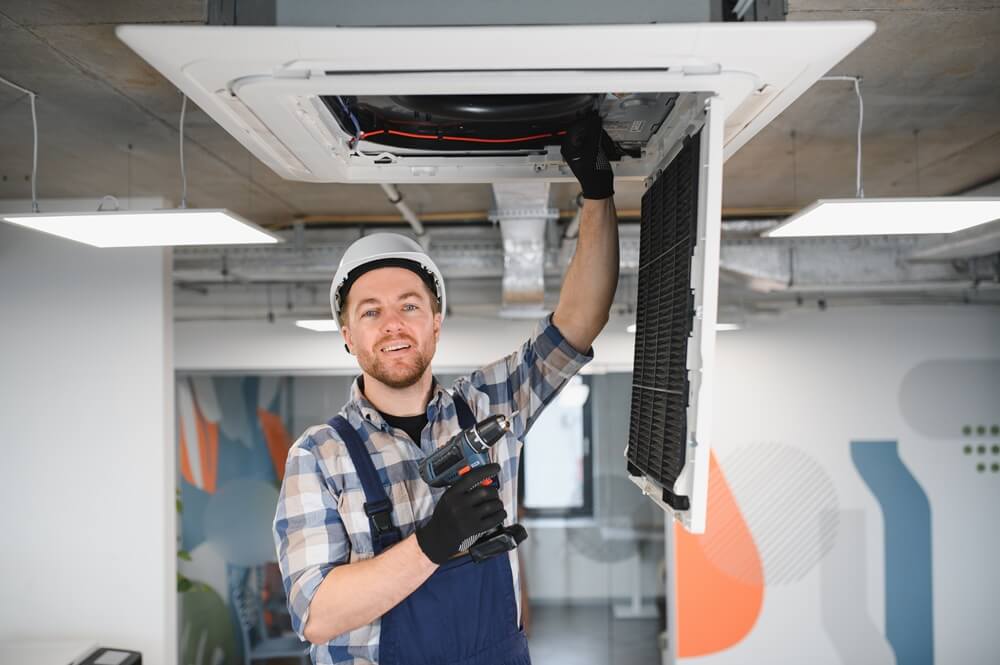 HVACR technician in overalls and hard hat servicing a commercial ceiling-mounted air conditioning unit.