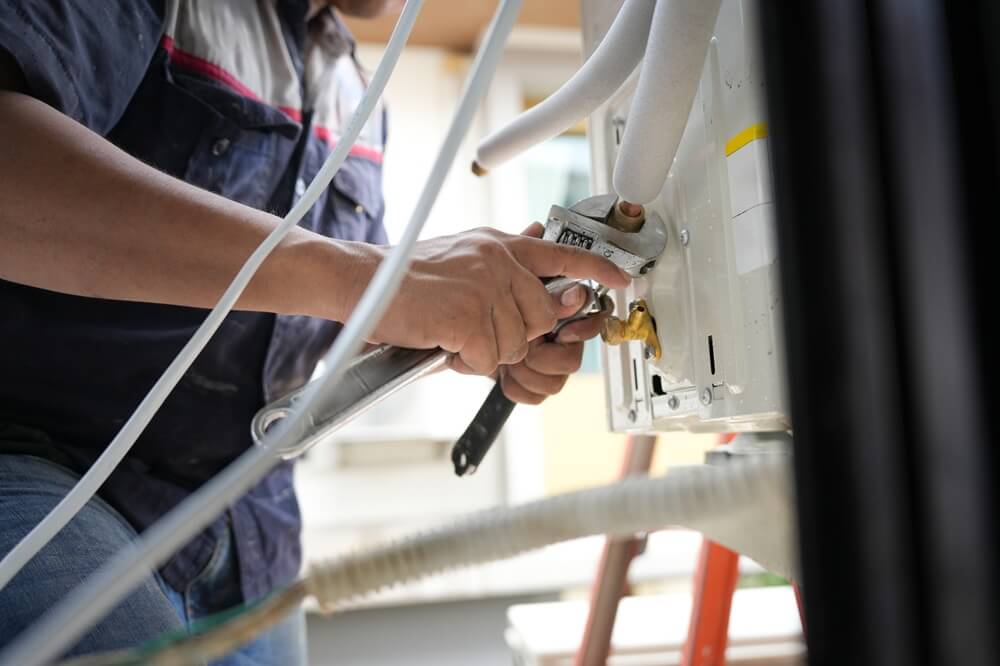 Technician tightening pipework on an outdoor air conditioning unit using a wrench during HVACR installation.