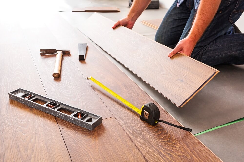 Contractor kneeling while positioning laminate flooring with tape measure and tools nearby.