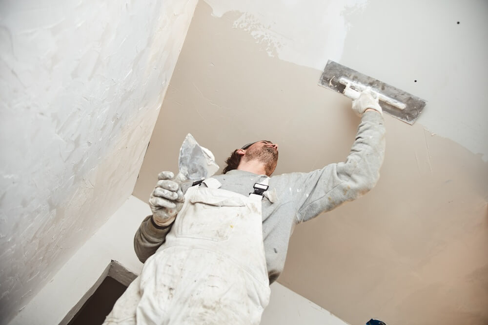 Plasterer in overalls applying plaster to a ceiling with a hawk and trowel, demonstrating expert internal finishing.