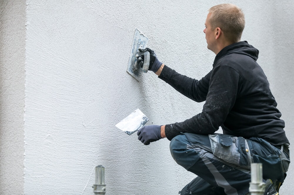Plasterer in dark workwear applying decorative plaster to an exterior wall using a trowel.