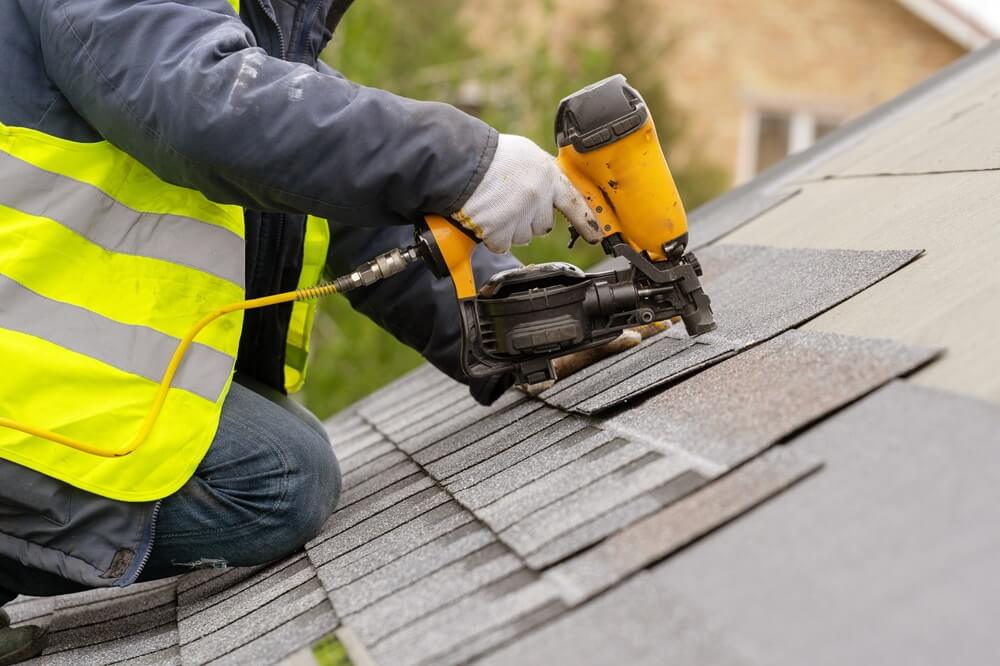 Roofer in hi-vis jacket using a nail gun to install asphalt shingles on a roof