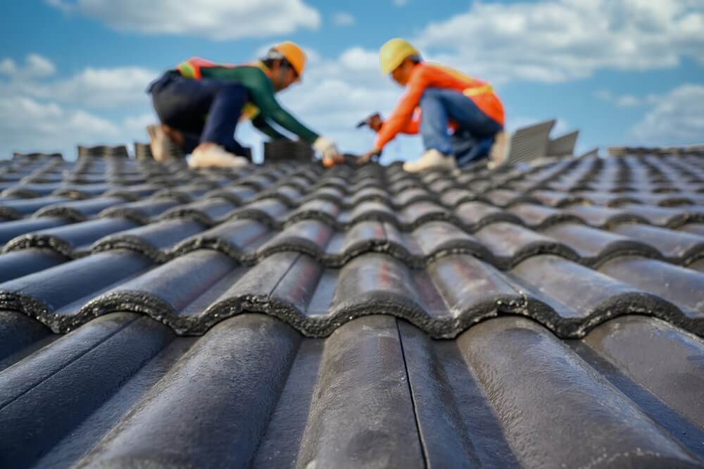 Two roofers installing black ceramic tiles on a pitched roof under a blue sky