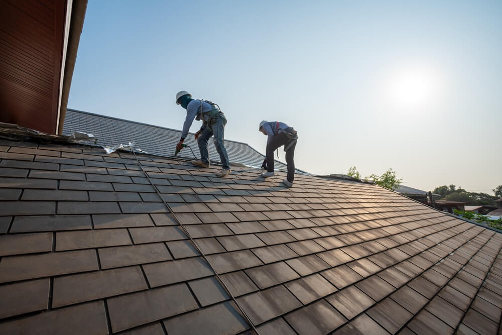 Two roofers installing slate-effect tiles on a large pitched roof under a clear sky