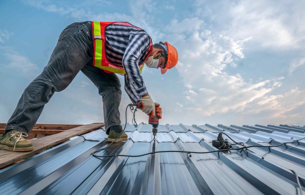 Roofer in PPE using a drill to install a metal roof under blue sky conditions