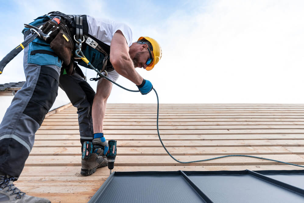 Roofer in safety gear fitting a panel to timber roof decking with a power drill