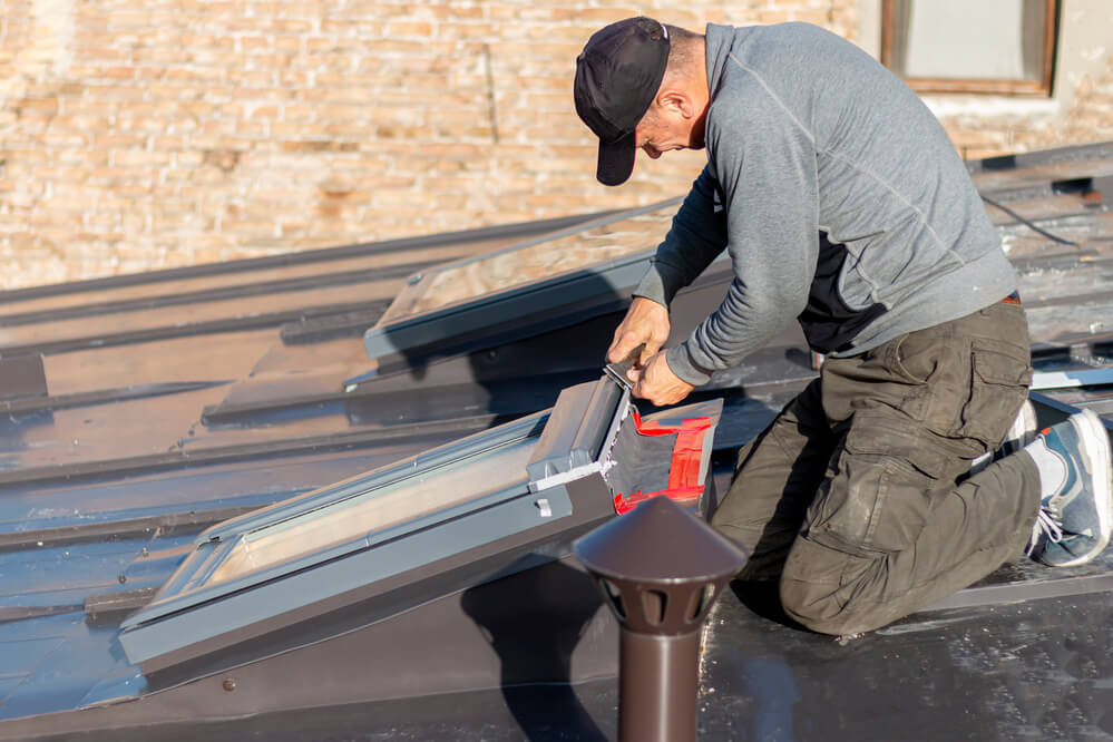 Roofer kneeling while fitting a skylight window into a flat roof using tools
