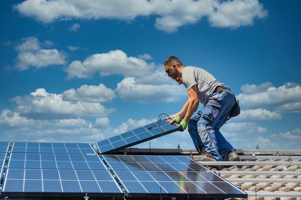 Two solar installers fitting PV panels on a tiled roof beneath clear blue skies.