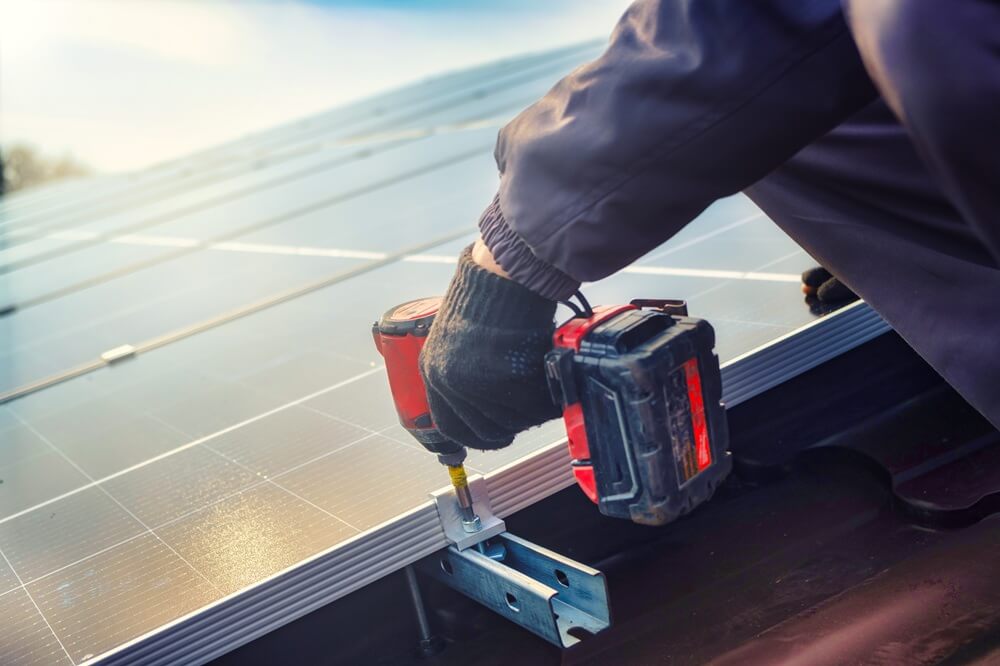 Close-up of a tradesperson using a cordless drill to fix a solar panel to a mounting rail.