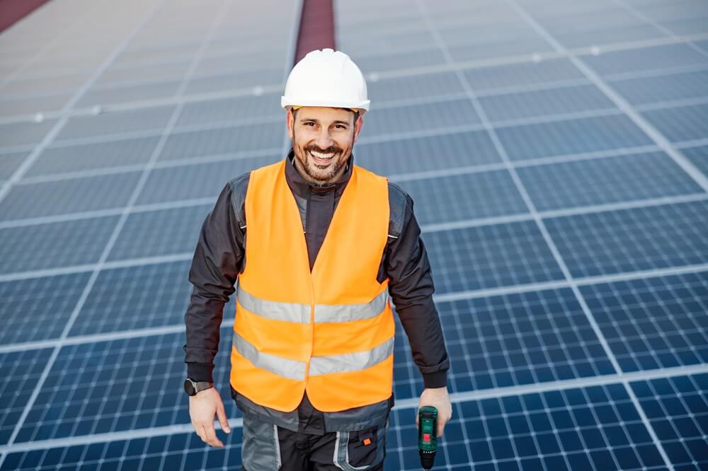 Smiling solar panel installer standing on a completed PV system, holding a drill.