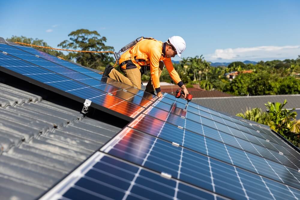 Installer drilling rooftop solar panels onto a metal roof in a suburban area.