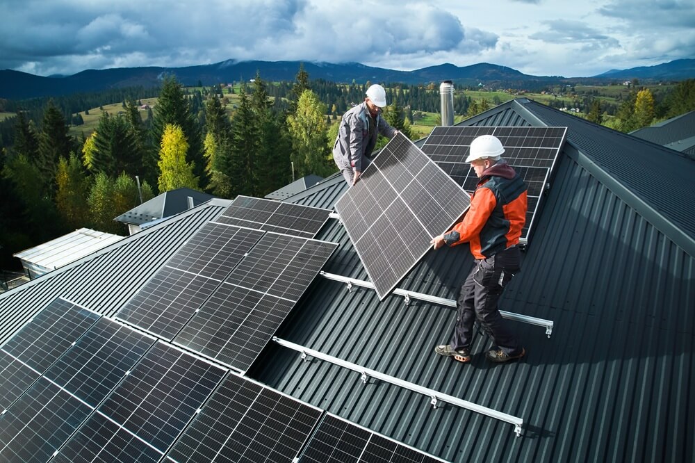 Two solar installers fitting PV panels on a pitched metal roof in a rural area.