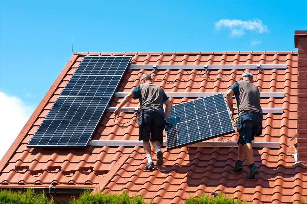 Two tradesmen installing solar panels on a red tiled residential roof in bright weather.