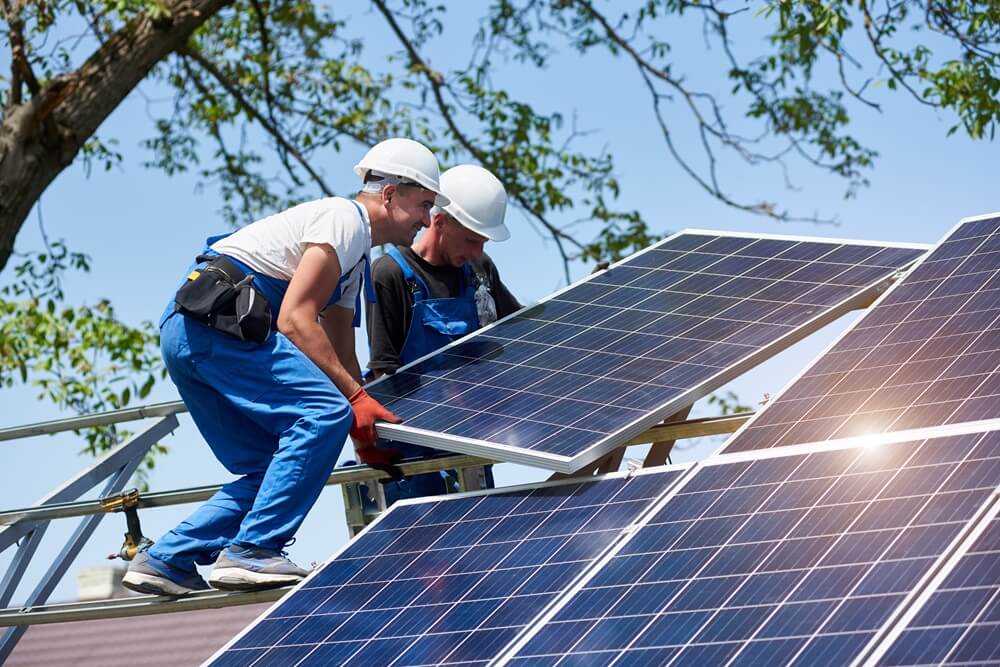 Two solar panel installers in hard hats mounting a panel onto a pitched roof during daytime.