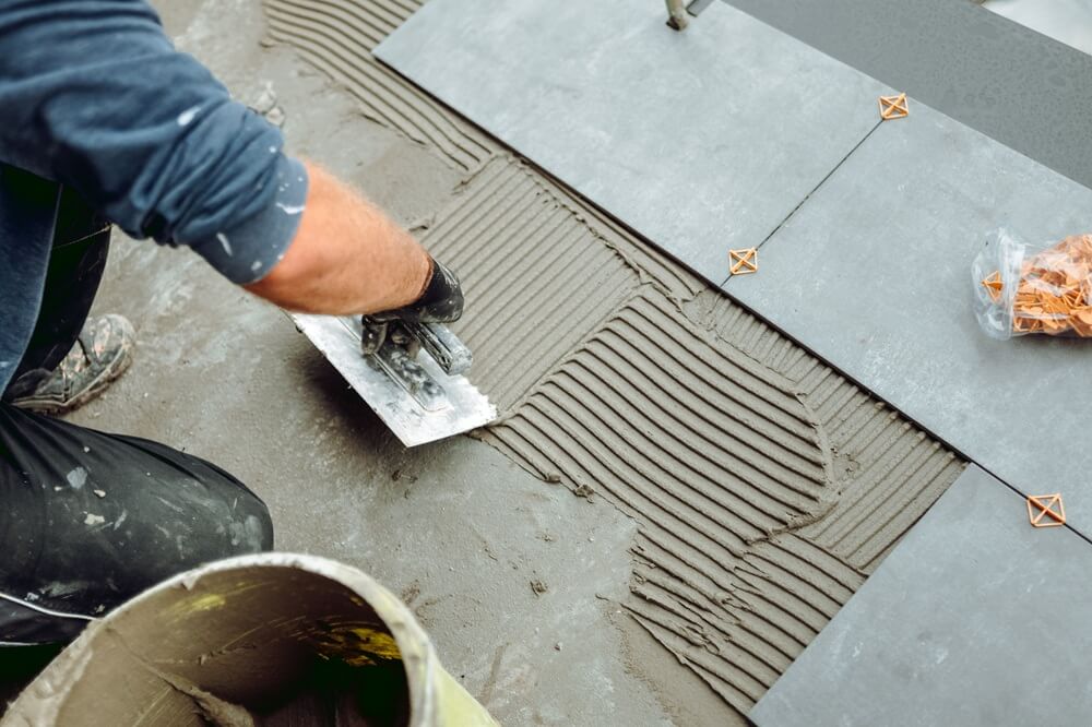 Flooring contractor spreading adhesive with a notched trowel before laying ceramic tiles.