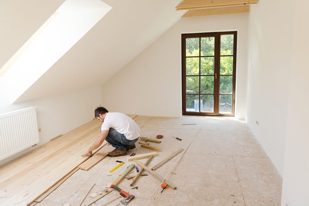 Worker installing wooden floorboards in a converted attic space, surrounded by tools and timber materials.